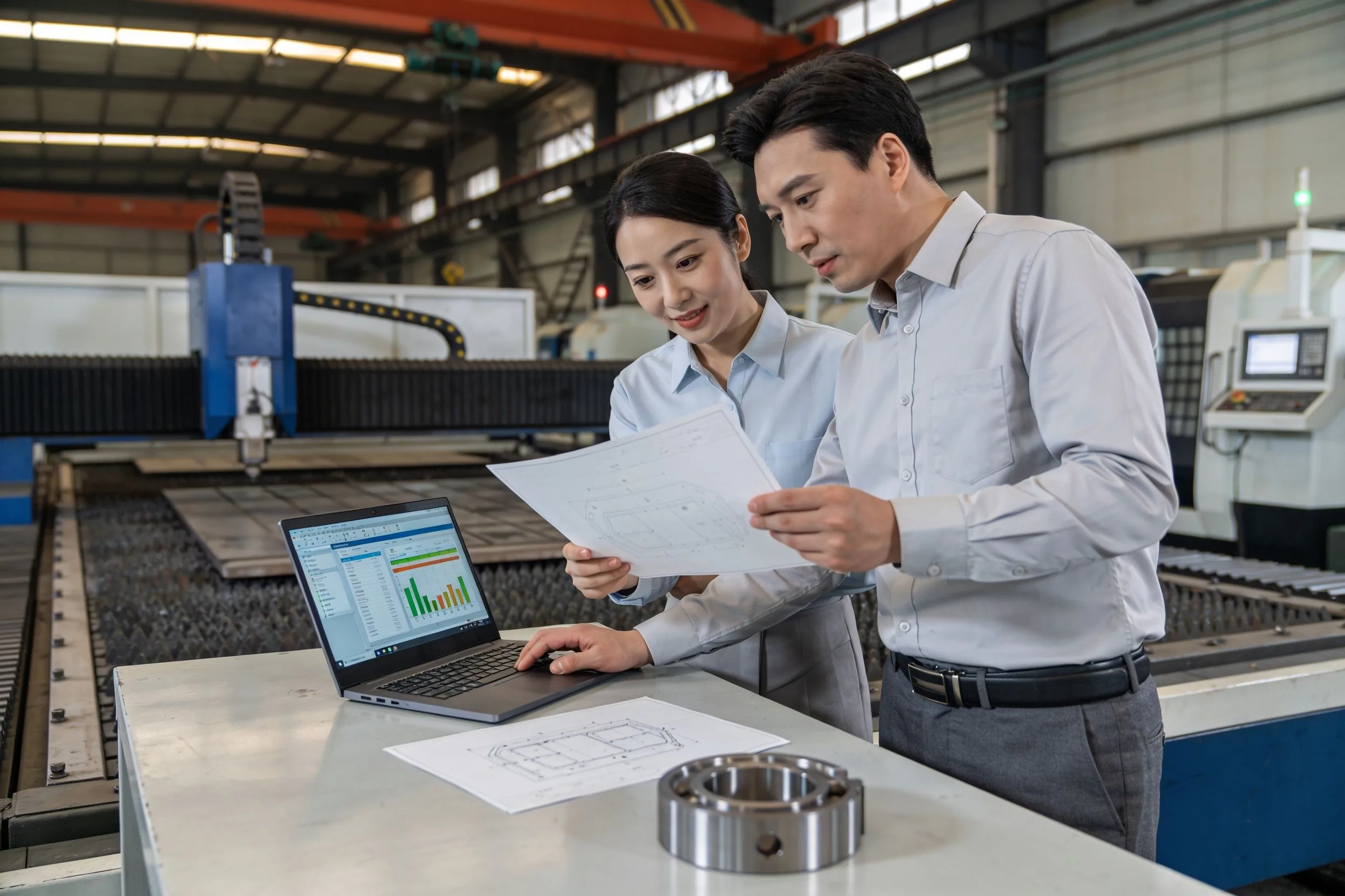 Commercial and production colleagues reviewing a component drawing beside a manufacturing line